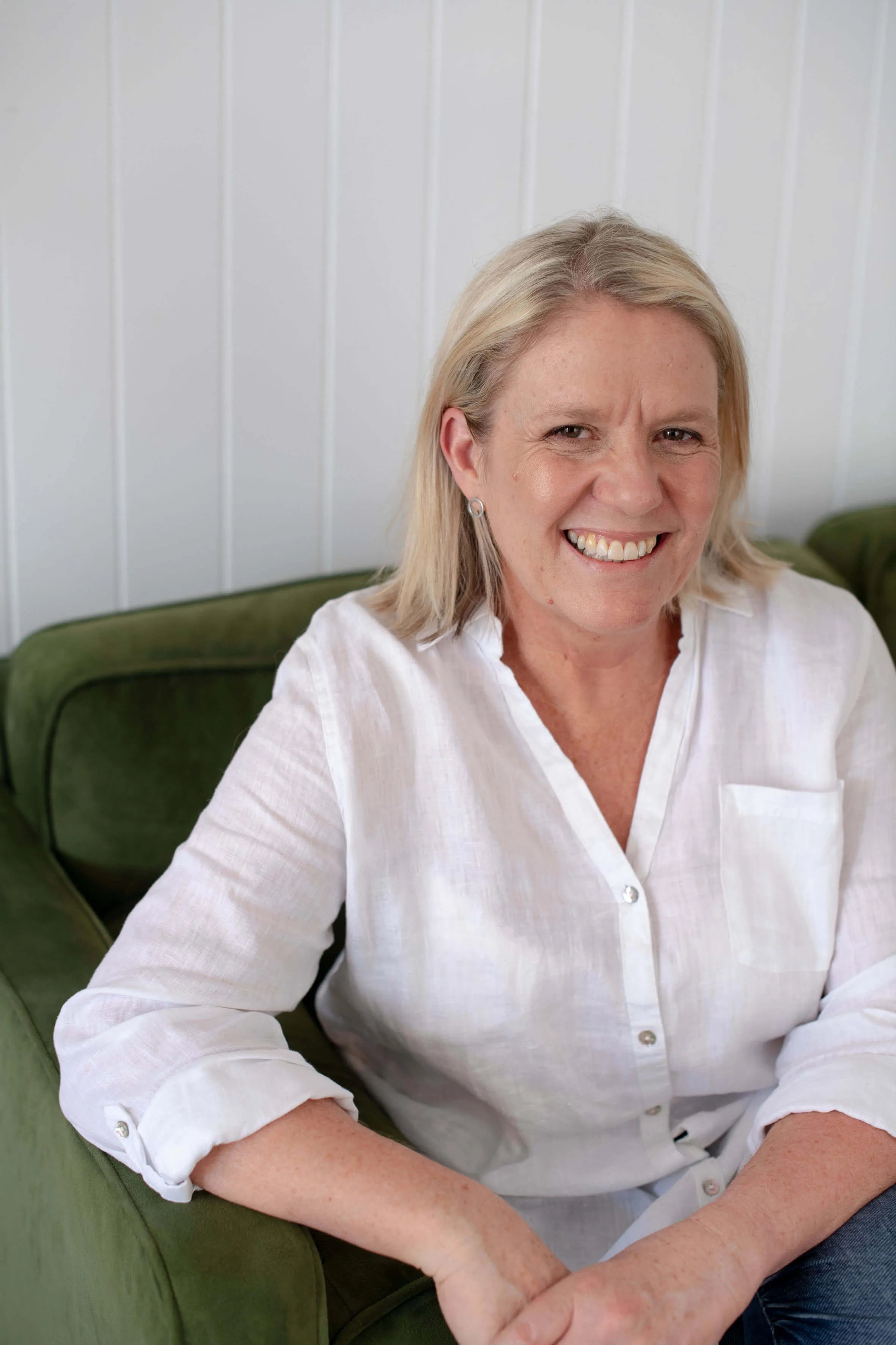 Marketday founder Michelle Hildebrand.  A smiling blonde woman wearing a white linen button-up shirt and blue jeans, seated on an olive green armchair against a white paneled wall.