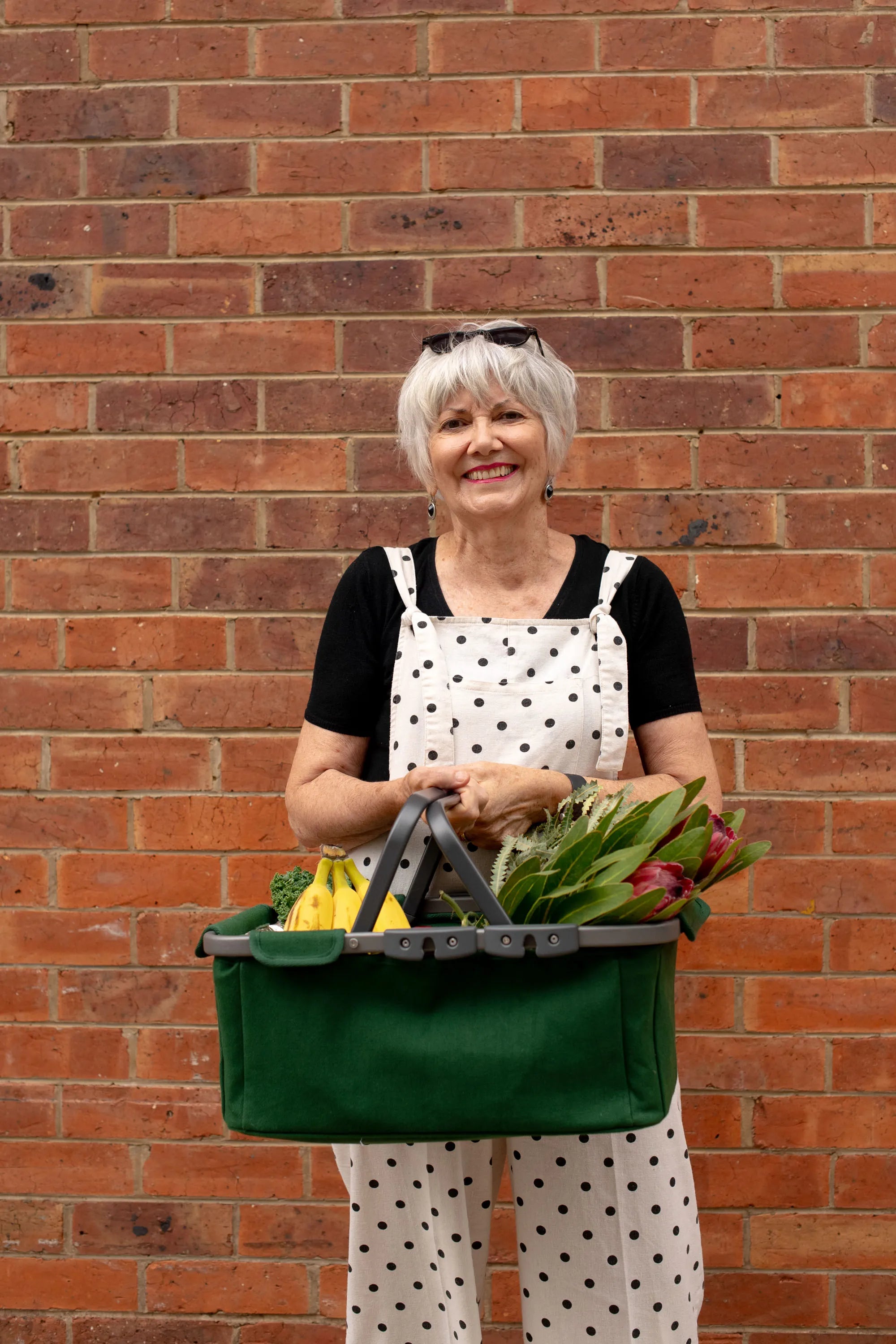 A smiling woman with short gray hair stands in front of a red brick wall, holding a green fabric produce basket filled with fresh vegetables. She is wearing a black top, white overalls with black polka dots, and sunglasses are resting on her head.